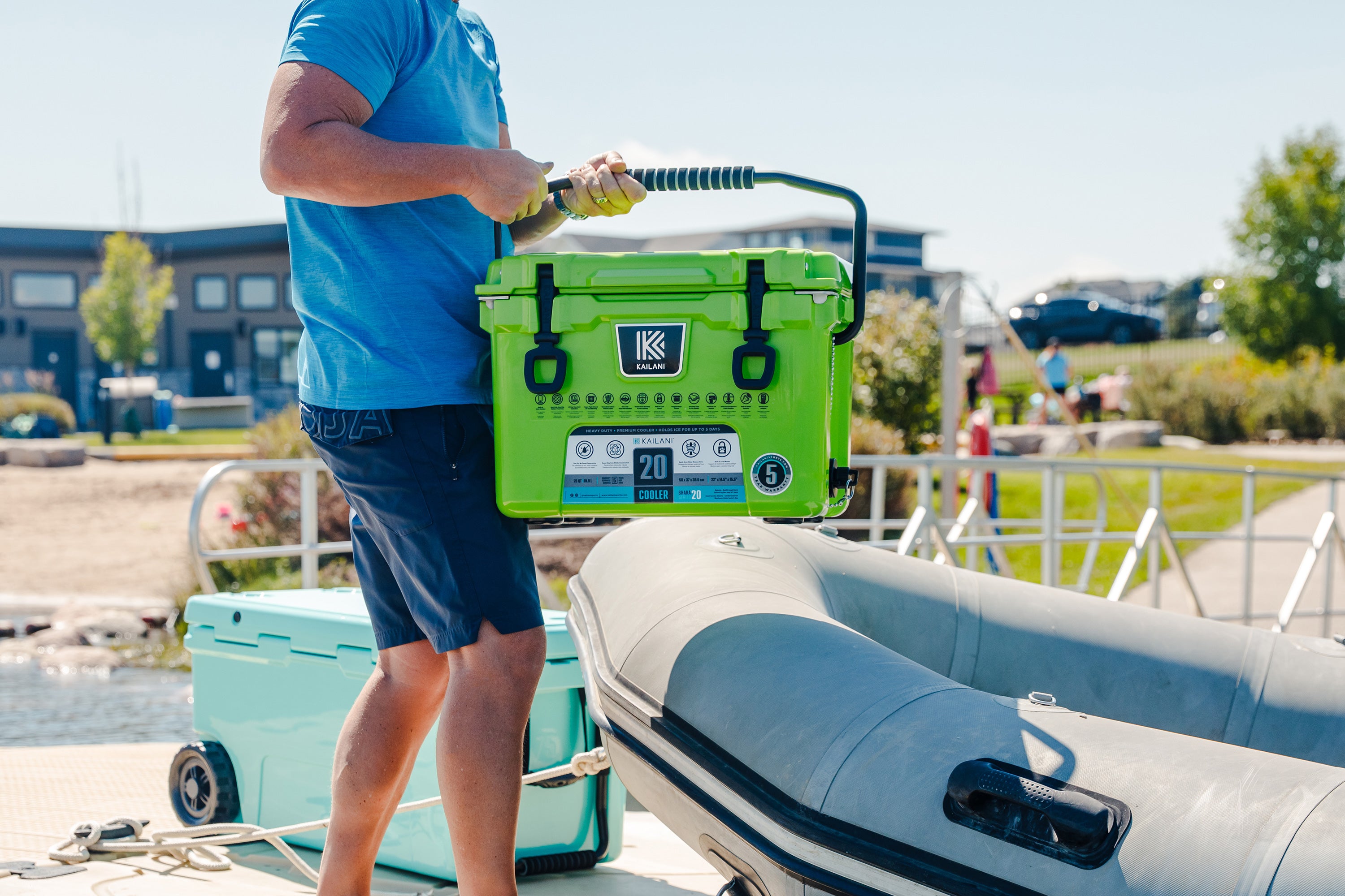 Person holding a green cooler box near an inflatable boat on a sunny day.